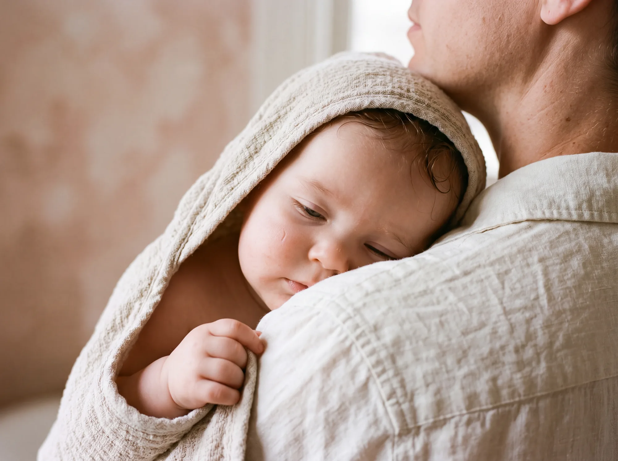A sleepy baby wrapped in a hooded towel resting on a parent’s shoulder after the bath.